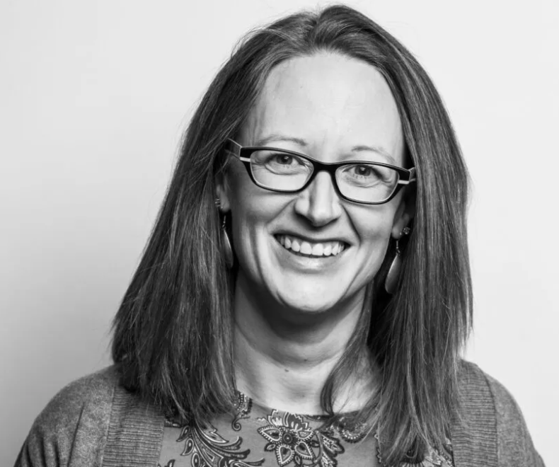 Smiling woman with long straight hair and glasses, wearing a patterned top and earrings, in a black and white portrait.